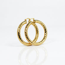 A pair of 18K gold hoop earrings with a textured, pixelated or tiled surface finish, displayed on a clean white reflective surface.