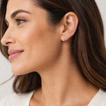 A close-up side profile of a woman wearing a small gold cross stud earring with tiny clear stones, set against a clean light gray background.