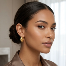A close-up portrait of a woman wearing a textured gold crescent-shaped hoop earring, set against a softly blurred indoor background.