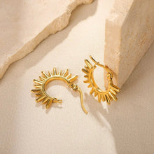 A pair of gold hoop earrings with a sunburst or flame-like radiating edge, displayed on a textured beige fabric next to rough stone blocks.