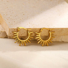 A pair of gold hoop earrings with a sunburst design, resting on a textured beige stone slab.