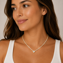 A woman wearing a rhinestone chain necklace with a small heart-shaped crystal pendant, styled with a white ribbed tank top against a neutral light beige background.