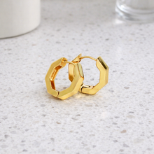 A pair of gold geometric hoop earrings with a faceted, multi-sided design, resting on a light speckled countertop.