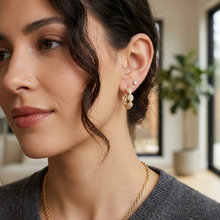 A close-up of a woman wearing a delicate gold huggie hoop earring with small clear stones, paired with a matching gold chain necklace. The background shows a softly blurred indoor setting with a plant.
