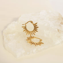 A pair of gold-toned hoop earrings with a sunburst or flame-like radiating design, displayed on a white textured stone against a warm cream background.