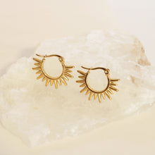 A pair of gold-toned hoop earrings with a sunburst or flame-like radiating design, displayed on a textured white stone against a warm cream background.