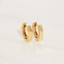 A pair of gold huggie hoop earrings with a modern, faceted geometric design, displayed on a clean white reflective surface.