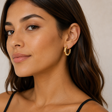 A close-up of a woman wearing a small gold geometric hoop earring, shown from the chest up against a soft beige background.