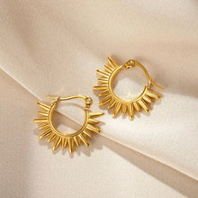 A pair of gold hoop earrings with a sunburst or starburst radiating from the outer edge, displayed on a draped beige fabric background.