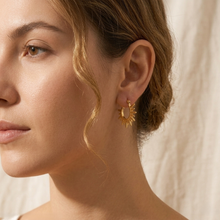A close-up side profile of a woman wearing a small gold sunburst hoop earring, set against a soft beige textured background.